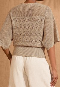 Kobieta w beżowym swetrze z dzianiny z wzorem w zygzak i siateczkowym, nosząca luźne kremowe spodnie, pokazana od tyłu na brązowym tle.