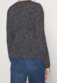 Haut noir avec un motif de cœur blanc, manches longues et ourlet peplum. Le tissu semble léger avec une légère texture. Porté avec un jean bleu.