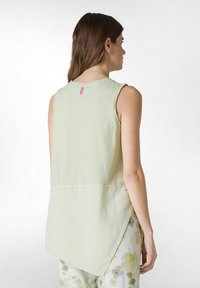 Top sin mangas de color verde claro, hecho de una tela suave, con un dobladillo asimétrico y una pequeña etiqueta rosa en la parte trasera. Diseño simple y minimalista.