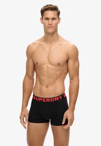 Boxer-briefs noirs avec une large ceinture rouge portant le logo "SUPERDRY". Le tissu semble lisse et ajusté. Modèle se tenant dans une pose neutre.