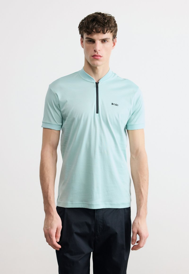 Νεαρός άνδρας που φοράει ανοιχτό γαλάζιο κοντομάνικο πουκάμισο με φερμουάρ μπροστά και μαύρο παντελόνι, στέκεται μπροστά σε απλό λευκό φόντο.