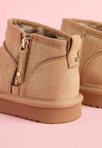 Botas tobilleras en oro rosa, hechas de suave gamuza, con cierre de cremallera y suela texturizada. Forradas con material acolchado para mayor comodidad.