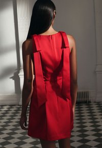 Červené šaty s přiléhavým živůtkem, širokými ramenními popruhy zdobenými mašlemi a skrytým zipem na zádech. Hladký, saténový vzhled.