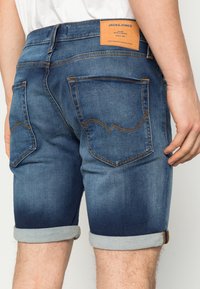 Muž nosí vyhrnuté tmavě modré džíny šortky se dvěma zadními kapsami a hnědou nášivkou značky Jack & Jones.