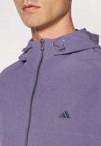 Lilla zip-up jakke med hette, snøring og brodert Adidas-logo. Glatt stoff med subtil tekstur.