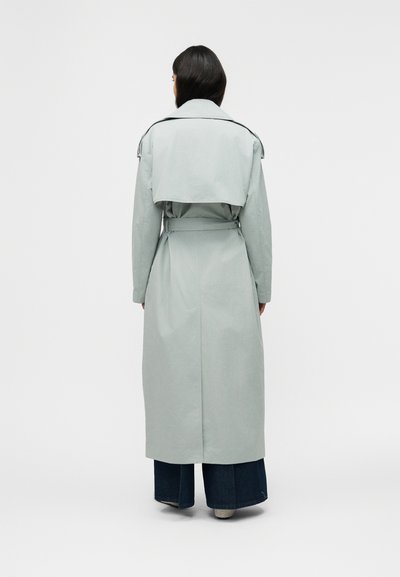 Γυναίκα με μακριά σκούρα μαλλιά που φοράει ένα ανοιχτό γκρι παλτό καμπαρντίνα με ζώνη και σκούρο τζιν φαρδύ κάτω μέρος, στέκεται πλάτη σε απλό φόντο.