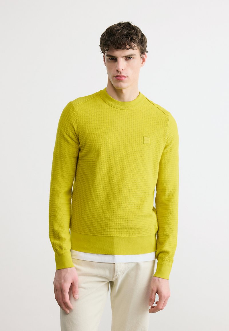 Jeune homme portant un pull texturé jaune vif par-dessus une chemise blanche et un pantalon beige clair, debout devant un fond blanc uni.