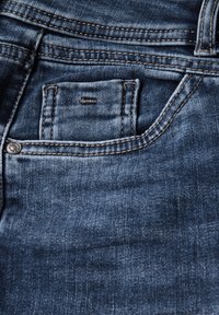 Los jeans de mezclilla azul oscuro presentan un diseño clásico, con costuras visibles y un pequeño bolsillo frontal. La textura es suave con un ligero brillo.