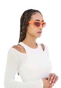 Occhiali da sole arancioni con montatura angolare e lenti rosse, indossati da un modello con capelli trecciati, abbinati a un top bianco con texture.