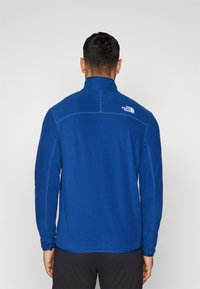 Man som bär en blå fleecetröja med "The North Face"-logotypen på övre högra ryggen, ståendes mot en enfärgad ljus bakgrund.