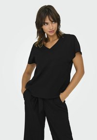Černý top s výstřihem do V a krátkými rukávy, měkká látka, volný střih, zobrazeno s koordinujícími černými kalhotami. Minimalistický design bez viditelných vzorů.