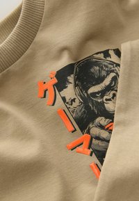 Beige pulóver, gorilla grafikával és merész narancssárga szöveggel. Az anyag sima tapintású, bordázott nyakkal.