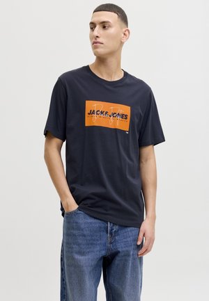 Młody mężczyzna w czarnej koszulce Jack & Jones z pomarańczowym logo i niebieskich dżinsach, stojący z jedną ręką w kieszeni, patrzący na bok.