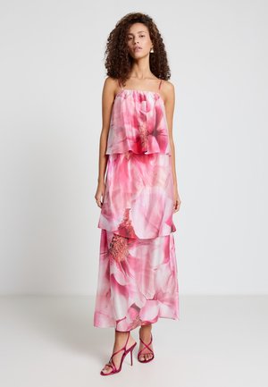 Femme portant une robe maxi sans manches à volants avec un imprimé floral rose, associée à des sandales à talons hauts roses avec des lanières, debout devant un fond uni.