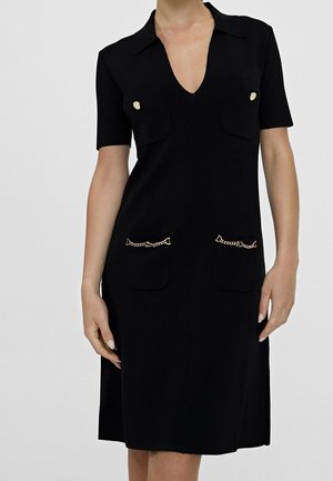 Schwarzes Kleid mit kurzen Ärmeln, Kragen, V-Ausschnitt und zwei Brusttaschen. Goldene Kettenakzente auf den vorderen Taschen. Glatte Stoffstruktur.