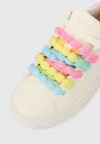 Fehér bőrsportcipő színes, bolyhos fűzőkkel, rózsaszín, sárga, kék és zöld színekben. A dizájnt lekerekített gyöngyök díszítik, amelyek a fűzők mentén helyezkednek el.