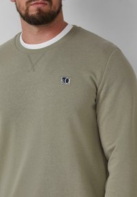 Uomo con barba che indossa un maglione girocollo di un leggero verde oliva con un piccolo logo quadrato "s.C" sul petto, sopra una t-shirt bianca.