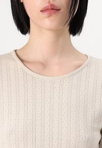Beige ribbet topp med en rund halslinje, med et teksturert mønster av vertikale linjer og små perforeringer, som fremhever det strikkede designet.