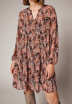 Femme portant une robe mi-longue à manches longues avec des motifs de feuilles orange, marron et blanches, assortie à des bottes beige montant jusqu'aux genoux.