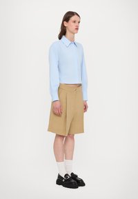 Helles blaues, kurz geschnittenes Hemd mit Kragen, kombiniert mit khakifarbenen, gefalteten Shorts und schwarzen Slippern. Weiße Socken runden das Outfit ab.