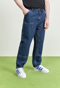 Jeans de mezclilla azul oscuro con un corte relajado, que presentan múltiples bolsillos y detalles de costura visibles. Combinados con zapatos blancos acentuados por rayas azules.