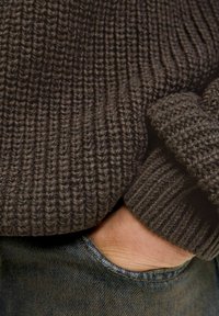 Pull en tricot marron avec des côtes texturées, poignets repliés, et une main reposant dans la poche d'un jean en denim bleu.