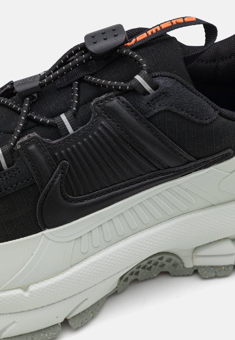 Czarne buty sportowe z teksturowaną cholewką z wypustkami, z miękką białą podeszwą i elastycznymi sznurowadłami zapewniającymi pewne dopasowanie. Jaskrawy pomarańczowy akcent.