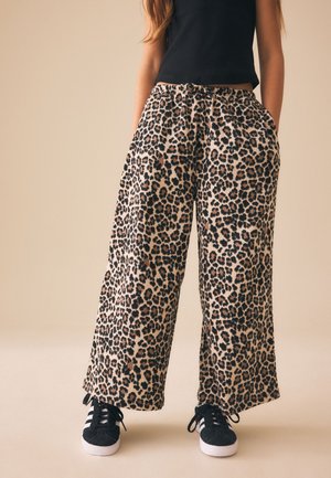 Kvinde iført leopardmønstrede bukser med bred benvidde, sort tanktop og sorte sneakers med hvide striber, stående med hænderne i lommerne.