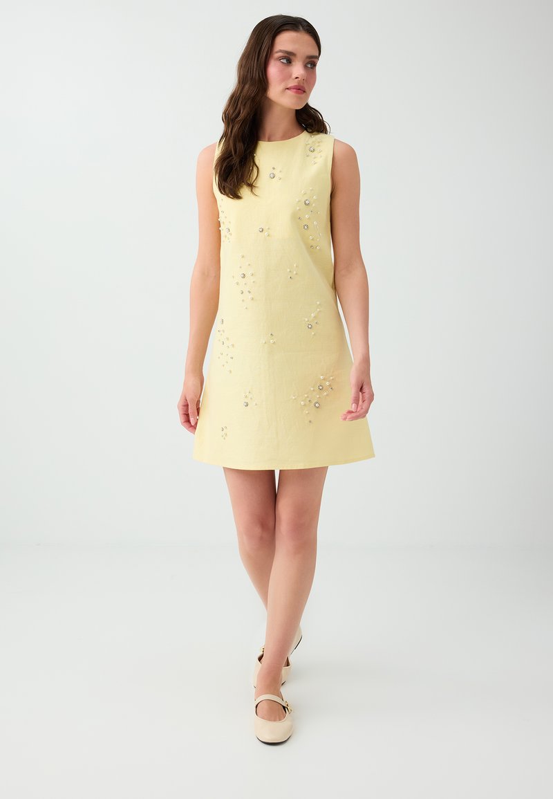 Femme aux longs cheveux bruns portant une robe sans manches jaune pâle avec des décorations florales subtiles et des chaussures Mary Jane beiges, marchant en avant.