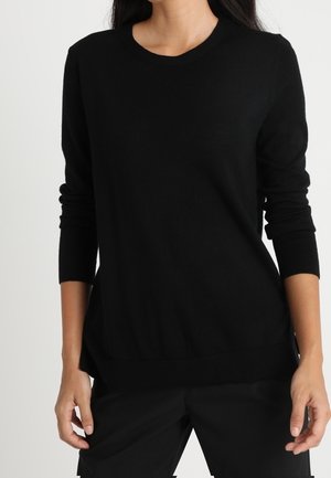 Žena oblečená v jednoduchém černém svetru s dlouhým rukávem a černých kalhotách, stojící na bílém pozadí.