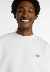 Felpa bianca con collo a girocolo a costine. Presenta un piccolo logo New Balance grigio sul lato sinistro del petto. Tessuto morbido, vestibilità casual, design minimale.
