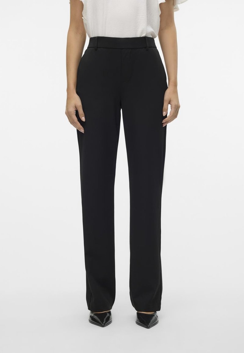 Femme portant un pantalon noir droit taille haute avec passants de ceinture, associé à un haut blanc et des chaussures noires à bouts pointus.