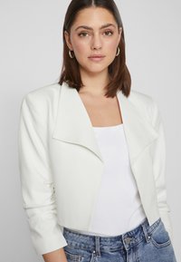 White cropped jacket with a wide collar, made of smooth material. Features rolled sleeves and a minimalist design, paired with a white top.