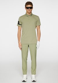 Olivgrön polo med svarta detaljer, kombinerad med matchande byxor. Modellen bär mörka solglasögon och en vit handske. Vita sneakers fulländar outfiten.