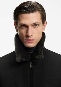 Junger Mann mit zurückgestrichenen Haaren, der einen schwarzen Mantel mit einem hohen, weichen Pelzkragen und einen schwarzen Rollkragenpullover vor einem einfarbigen Hintergrund trägt.