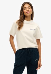 T-shirt en coton beige clair à manches courtes, avec le texte bleu "ATHLETICS" sur la poitrine, associé à un jogger en velours côtelé bleu foncé.
