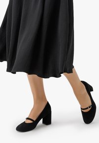 Melni zamšas apavi ar blokveida papēdi, ar noapaļotu purngali, ar šauru siksniņu pār pēdas vidu un teksturētu apdari.