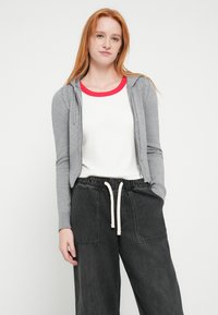 Γκρι ζακέτα με φερμουάρ και ριμπ μανσέτες και ραφές, φορεμένη πάνω από λευκό τοπ με κόκκινο γιακά, σε συνδυασμό με χαλαρό μαύρο παντελόνι με τσέπες.