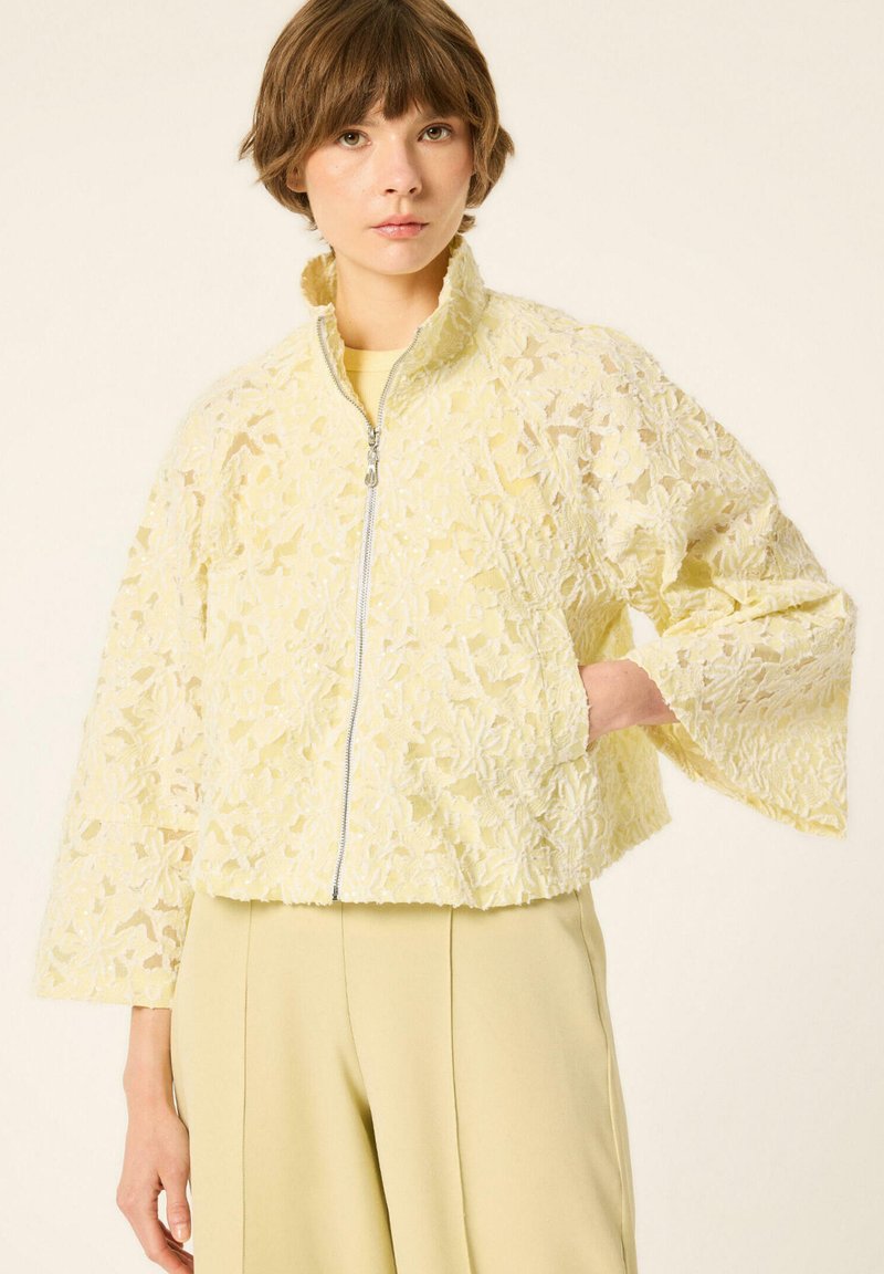 Jeune femme portant une veste zippée en dentelle fleurie jaune pâle et un pantalon taille haute assorti, main dans la poche.