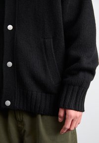 Cardigan nero lavorato a maglia con polsini e orlo a costine, dotato di bottoni a pressione argentati e una tasca laterale, abbinato a pantaloni verde oliva.