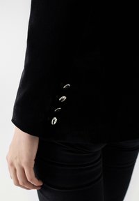Černý kordový blazer s klopovým límcem, zobrazující detail rukávu s čtyřmi stříbrnými knoflíkovými akcenty a hladkou texturou.