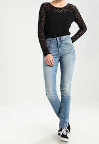 Schwarzes Langarmoberteil mit Spitze, kombiniert mit hellblauen Slim-Fit-Jeans und schwarzen Sneakern. Lässiges Outfit mit modernem Look.