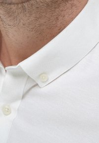 Chemise blanche boutonnée mettant en valeur un tissu texturé avec un col pointu et deux boutons blancs visibles au niveau du col. Vue en gros plan.