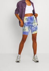 Tie-dye bike shorts in blue, yellow, and white. Made from a stretchy fabric. Paired with a white crop top and a purple jacket. Silver sneakers.