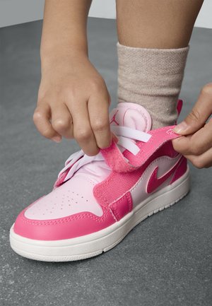 Les mains d'un enfant attachant une chaussure de sport rose et blanche avec une sangle auto-agrippante sur des lacets blancs, sur un sol gris, portant des chaussettes beiges.