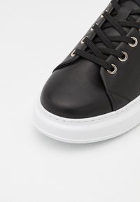 Black leather sneaker with a rounded toe, white rubber sole, and textured surface. Features metal eyelets and black laces.