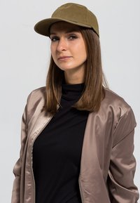 Gorra verde oliva con diseño estructurado, combinada con una chaqueta bomber satinada de color beige sobre un suéter negro de cuello alto. Texturas suaves y tonos neutros.