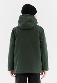 Groene waterdichte jas met een capuchon, featuring een gladde textuur, reguliere pasvorm en logo-detail op de achterkant. Zwarte geribbelde beanie gedragen.