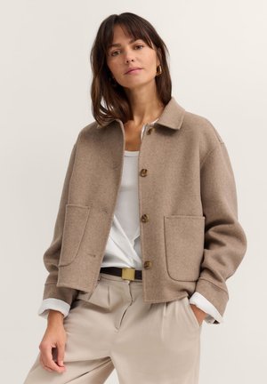 Frau trägt eine beige Wolljacke über einem weißen Hemd, beige Hosen mit einem schwarzen Gürtel, steht mit den Händen in den Taschen.