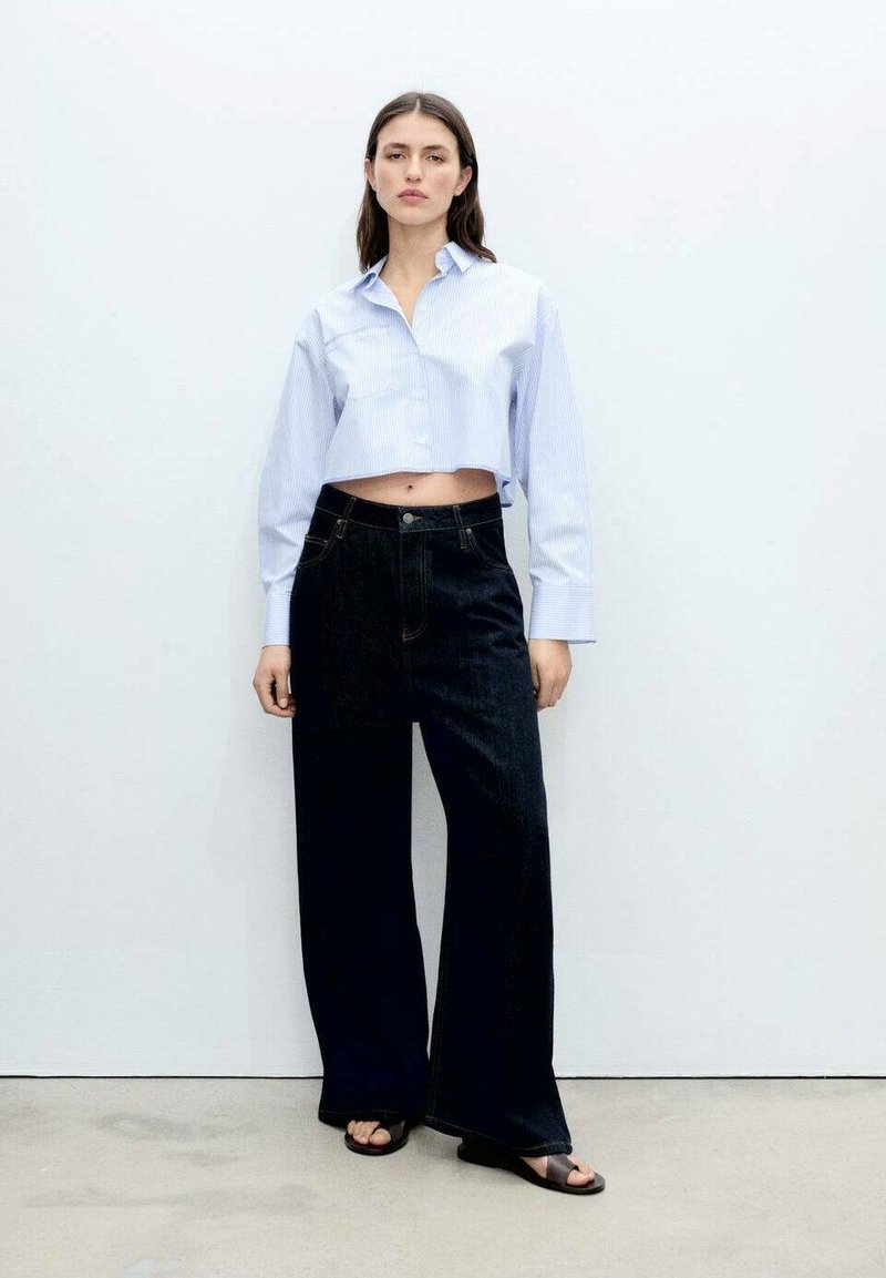Geknipte lichtblauwe gestreepte blouse, hoge taille donkere denim wijde jeans en zwarte sandalen, tegen een effen witte achtergrond.